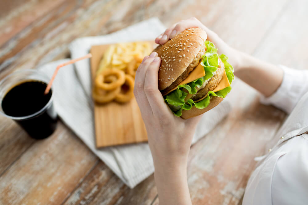 Ruky držiace hamburger, v pozadí s hranolkami, cibuľovými krúžkami a colou